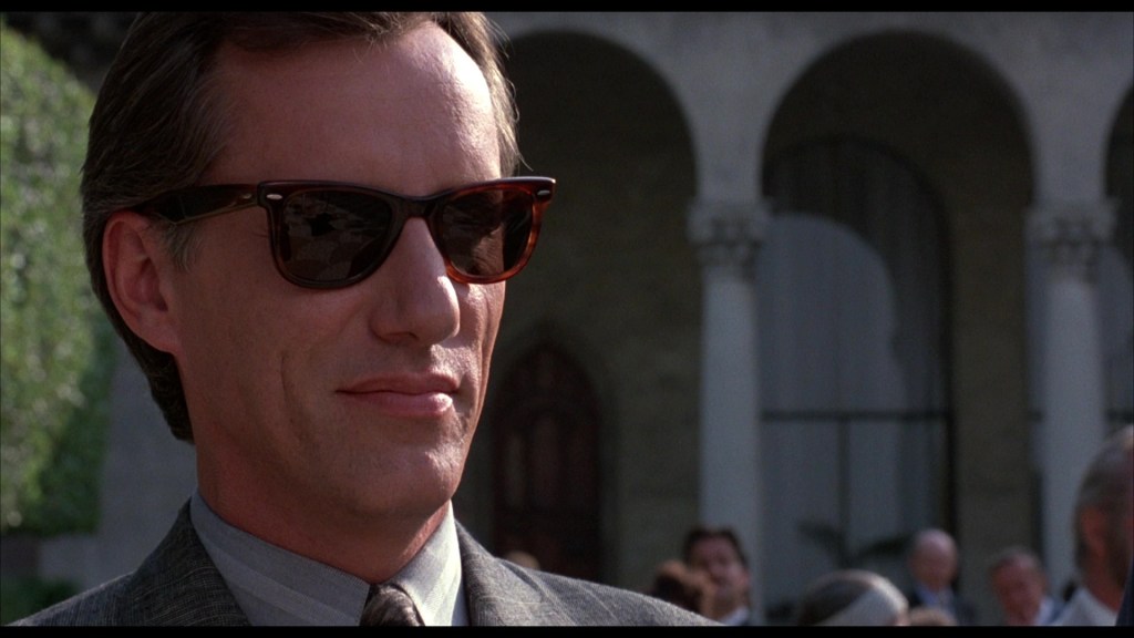 A man wearing sunglasses and a suit, with a serious expression, standing in front of a decorated building with arches.