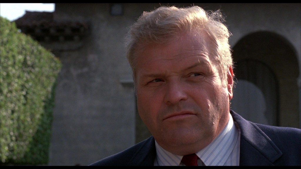 A man with blonde hair, wearing a suit and tie, is standing outside near a house, looking slightly discontent or skeptical.