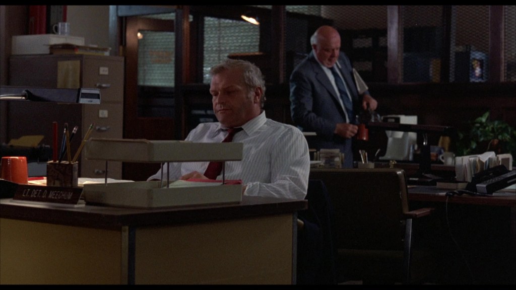 Two men in an office setting, one seated at a desk with a nameplate reading 'Lt. Det. D. Meechum' and the other standing with a coffee pot, surrounded by office supplies and furniture.