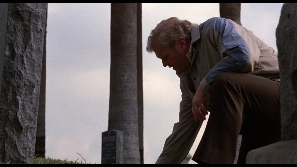 A man kneels beside a grave, gently touching the ground, with palm trees in the background.