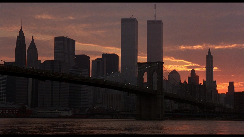 Silhouette of a city skyline at sunset featuring the Brooklyn Bridge and tall buildings, including the Twin Towers.