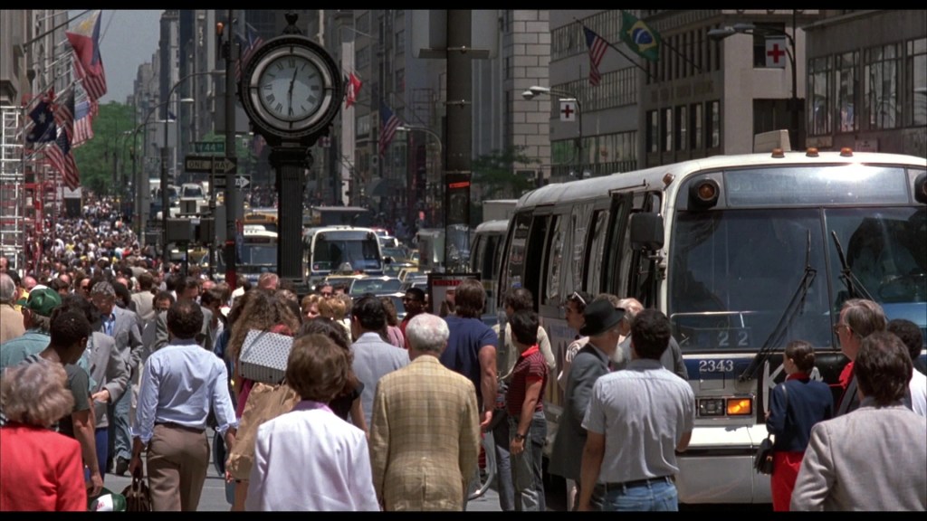 A busy city street filled with pedestrians, vehicles, and a large clock. American flags are displayed along the buildings, creating a vibrant urban atmosphere.