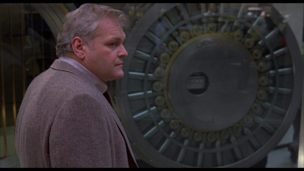 A man with short light hair and a serious expression stands beside a large bank vault door.