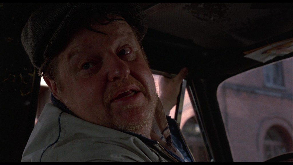 A middle-aged man with facial hair and wearing a flat cap, looking back from the driver's seat of a vehicle, appearing engaged in conversation.