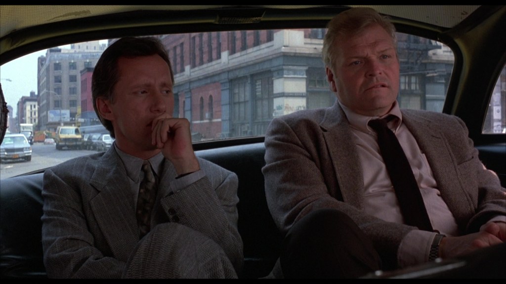 Two men sitting inside a taxi, looking contemplative with city streets visible through the window.