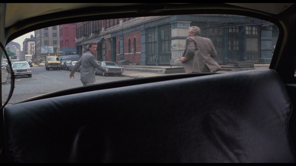 View from inside a car showing two men running on a street, with buildings and parked cars in the background.