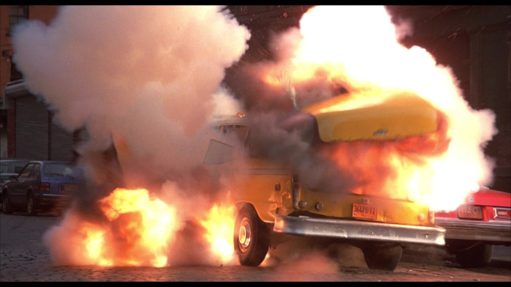 A yellow taxi cab explodes in a cloud of smoke and flames on a city street.