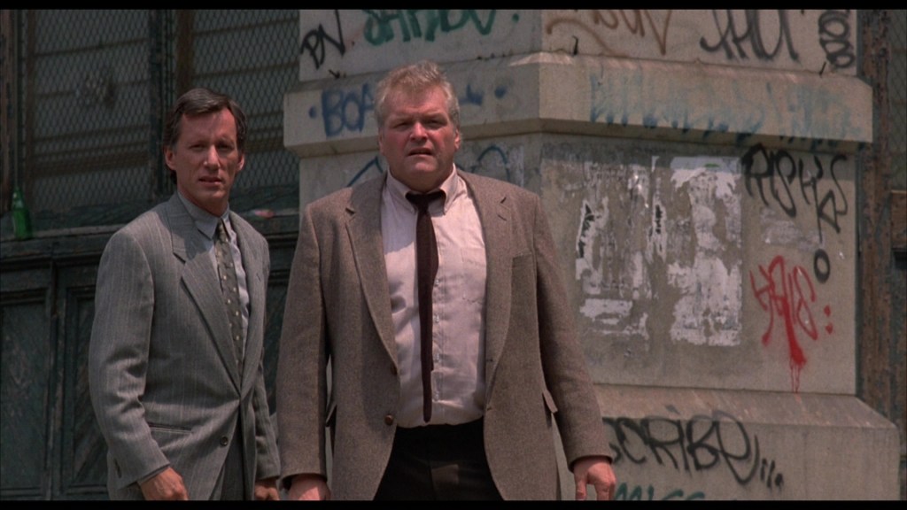 Two men in suits stand outside, looking surprised or concerned in front of a graffiti-covered wall.