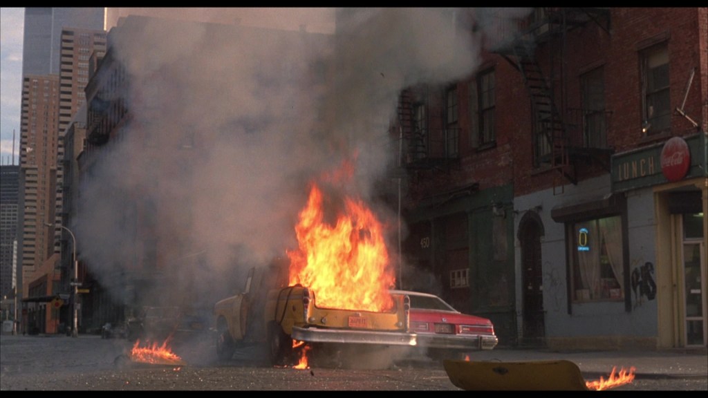 A street scene featuring a burning yellow taxi and a red car with smoke and flames, set against a backdrop of urban buildings.
