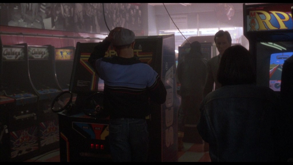 A dimly lit arcade with several vintage arcade machines, featuring players engaged with the games. A person in a striped sweater stands at one of the machines, while others are visible in the background.