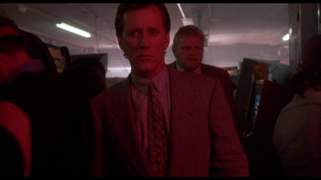 A man in a suit, looking concerned, walks through a crowded arcade with glowing lights and arcade machines in the background.