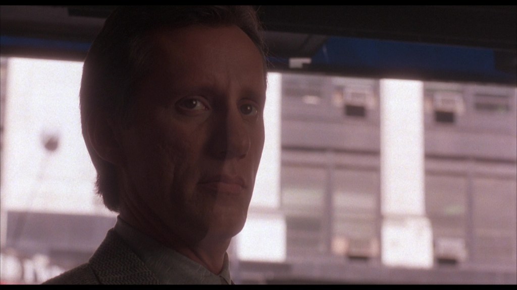Close-up of a man with short dark hair and a serious expression, set against a blurred city background.
