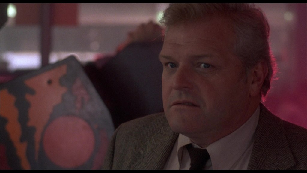 A close-up of a middle-aged man with a concerned expression, wearing a suit and tie, with a blurred colorful background.