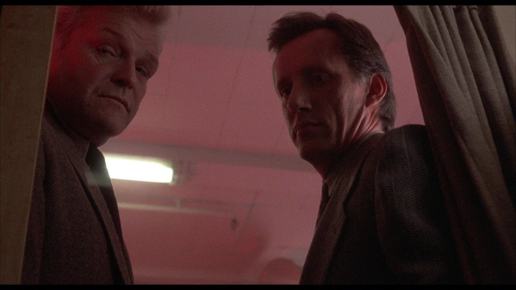 Two men looking intently towards the camera from behind a curtain, with a reddish light illuminating the scene.