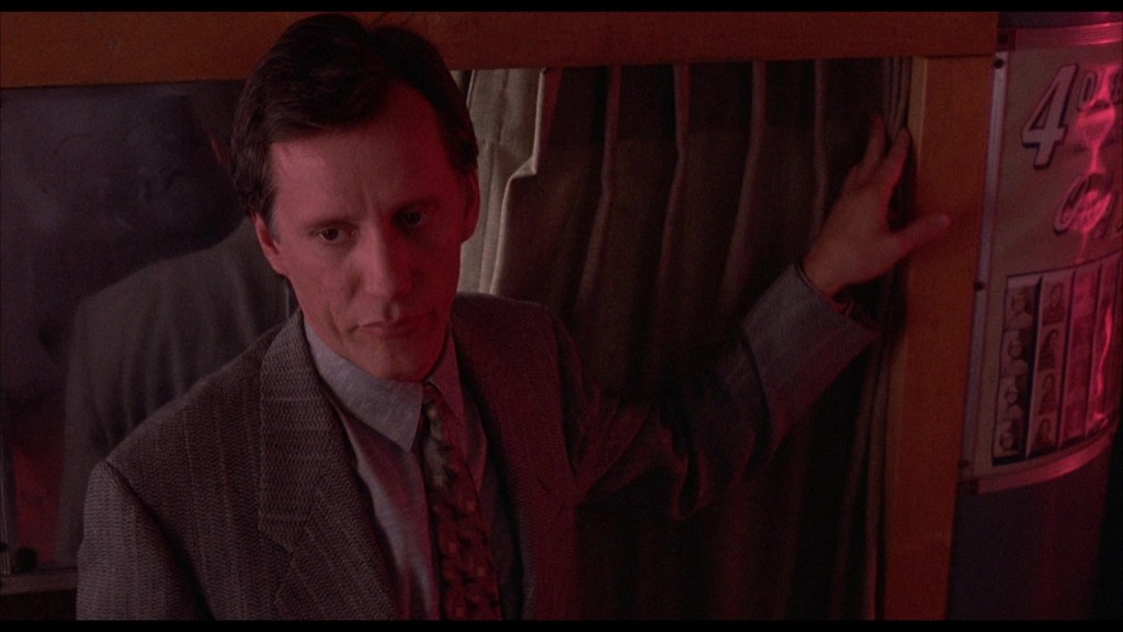 A man in a suit stands near a curtain, looking pensive. The scene is illuminated with a reddish hue, creating a dramatic atmosphere.