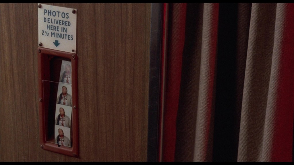 A photo booth with a sign reading 'PHOTOS DELIVERED HERE IN 2½ MINUTES' above a slot containing several photo strips.