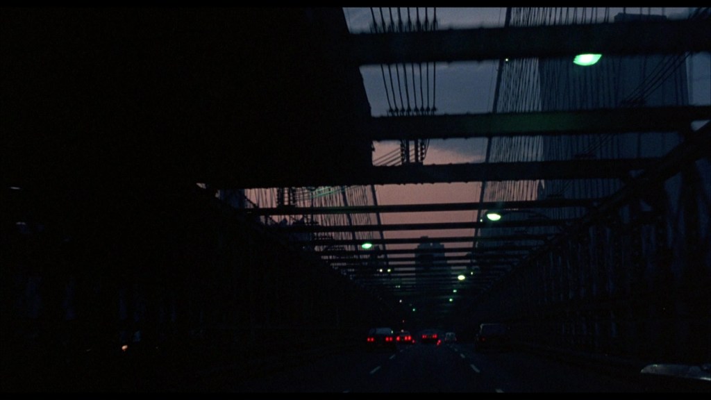 View of a darkened bridge roadway at dusk, with cars traveling underneath a lattice of beams and streetlights glowing in the darkness.