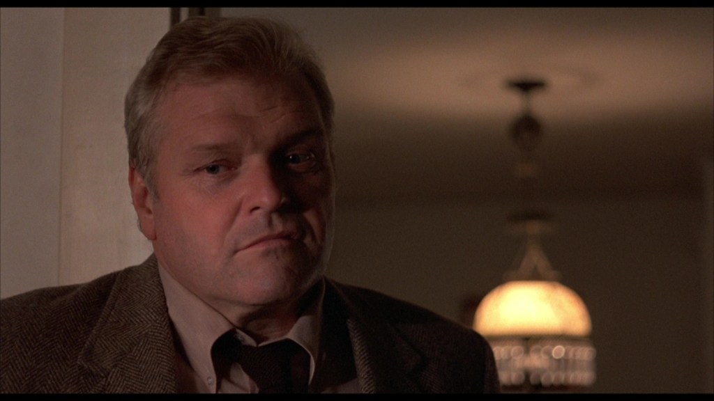Close-up of a middle-aged man with short, light hair, wearing a brown blazer over a light shirt, looking thoughtfully with a serious expression, background features a softly lit hanging lamp.