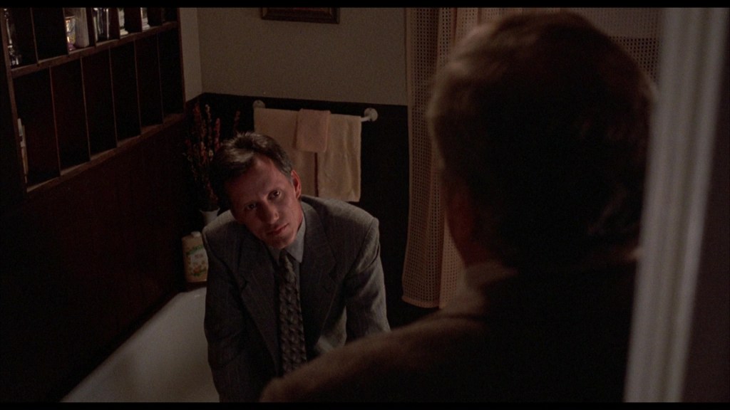 A man in a suit leans forward, looking intently at another man who is positioned out of focus in the foreground. The setting appears to be a bathroom, with a bathtub visible and towels hanging on the wall.