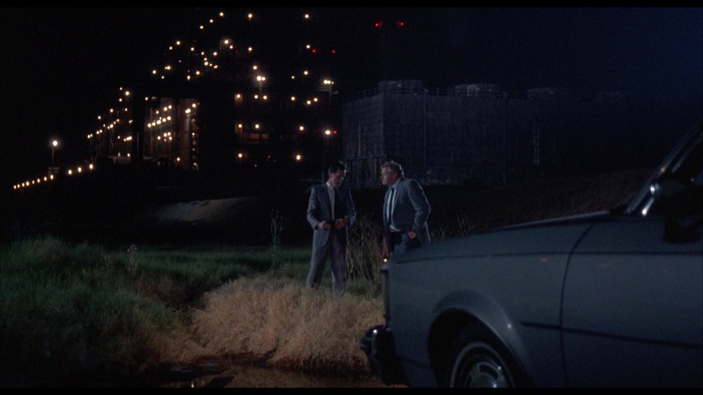 Two men in formal attire stand near a road at night, discussing while a gray car is parked nearby. In the background, a lit-up building is visible.