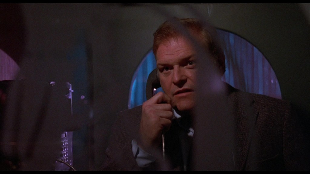 A man wearing a suit is speaking on the phone, with a concerned expression, partially obscured by a glass partition.