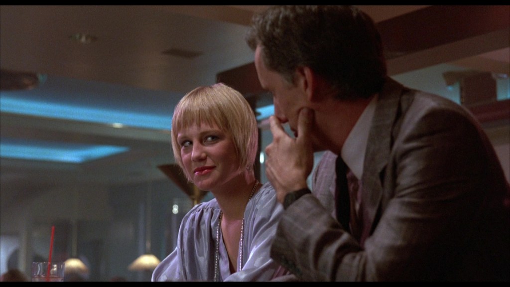 A woman with short blonde hair sits at a bar, smiling and engaging with a man in a suit, both appearing to have a conversation.