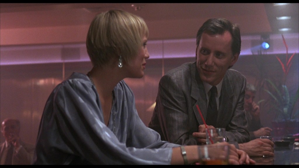 A man in a suit smiles at a woman with short hair and a stylish blue blouse, sitting at a bar with drinks in front of them. The atmosphere is dimly lit with a slight haze.