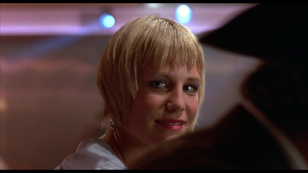 A close-up portrait of a woman with short, blonde hair and a slight smile, set against a softly illuminated background.