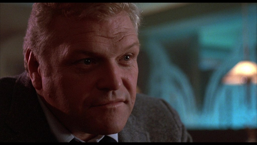 A close-up of a man with short light-colored hair and a serious expression, wearing a gray blazer, sitting in a dimly lit setting with blue and green background lighting.