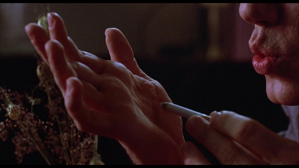 A close-up of a hand holding a pen, with another hand resting open nearby. The background features blurred flowers, creating an intimate and contemplative atmosphere.