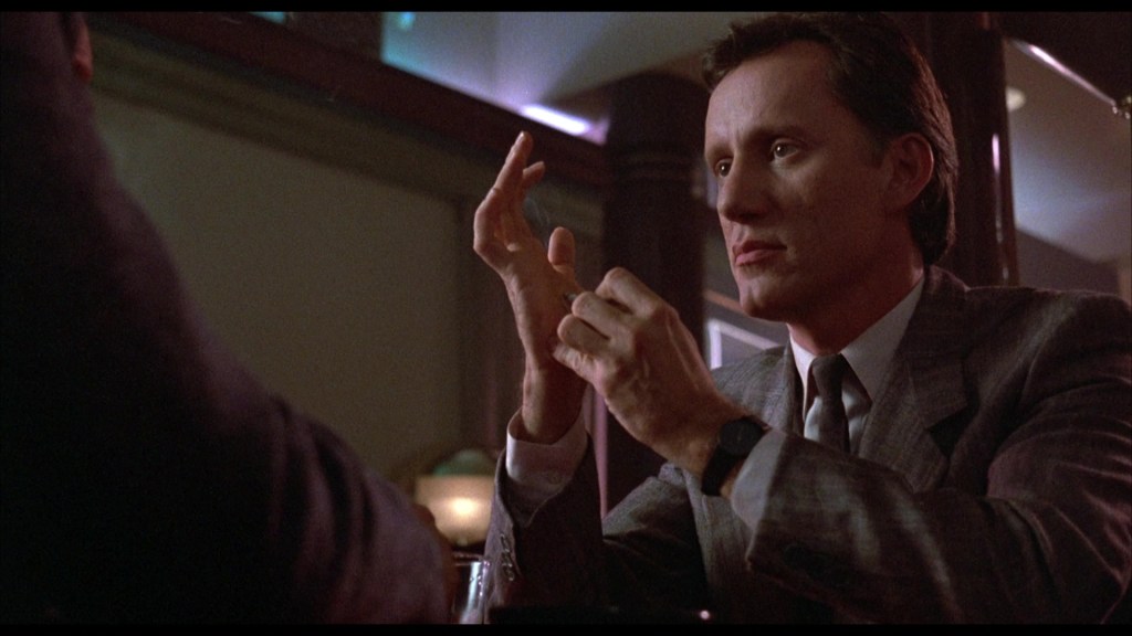 A man in a gray suit gestures while seated at a table, engaged in conversation. The background is softly lit, creating a dramatic atmosphere.