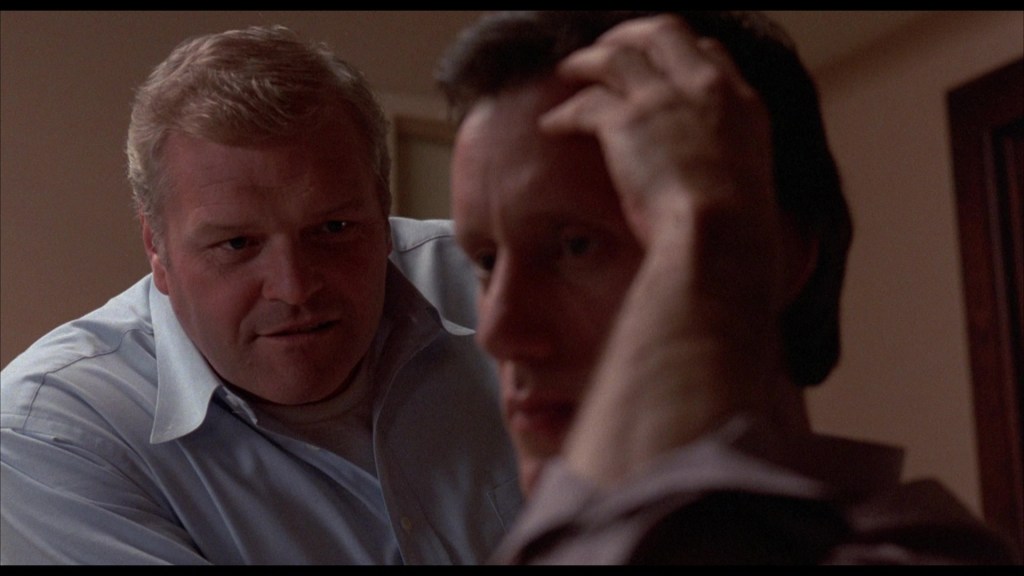 A close-up of two men in a tense conversation, with one man leaning in and looking closely at the other, who appears stressed and holds his head with his hand.