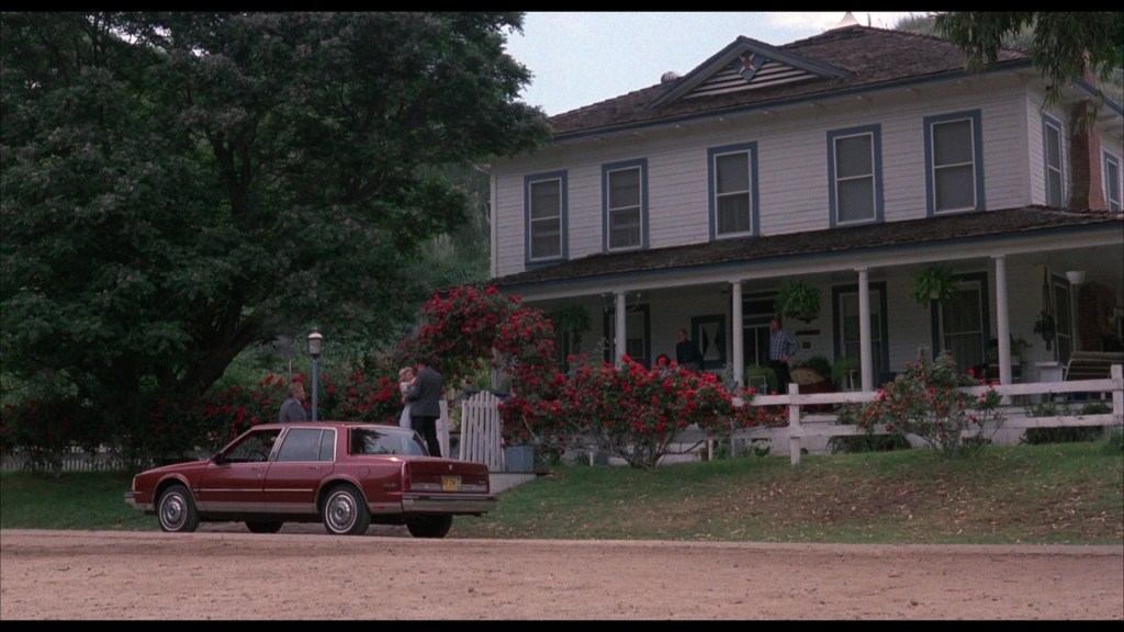 A red car parked in front of a large white house surrounded by greenery and colorful flowers, with people conversing on the porch.