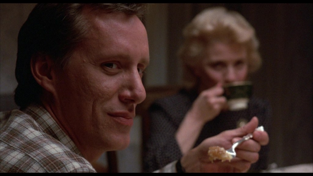 A man in a plaid shirt smiles slightly while holding a fork with food, as a woman in the background sips from a cup.