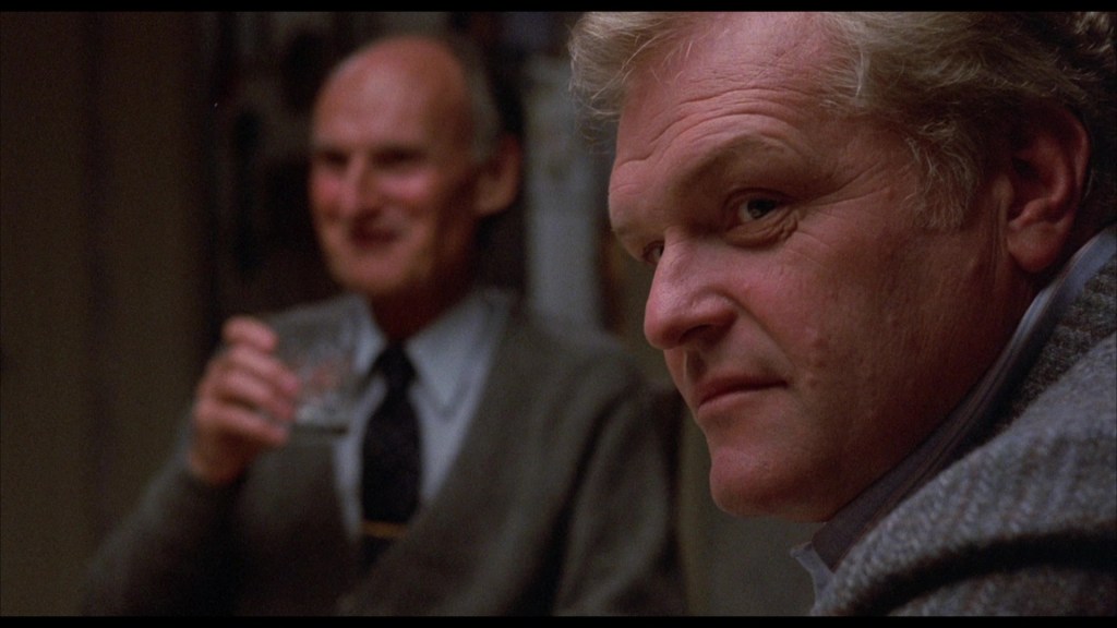 A close-up of a man with light hair and a serious expression, sitting at a table, while an older man in the background holds a glass and smiles.