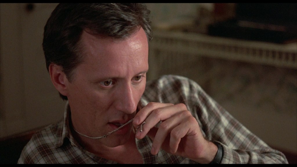 A man in a plaid shirt is thoughtfully holding a chain with a pendant, looking pensive.