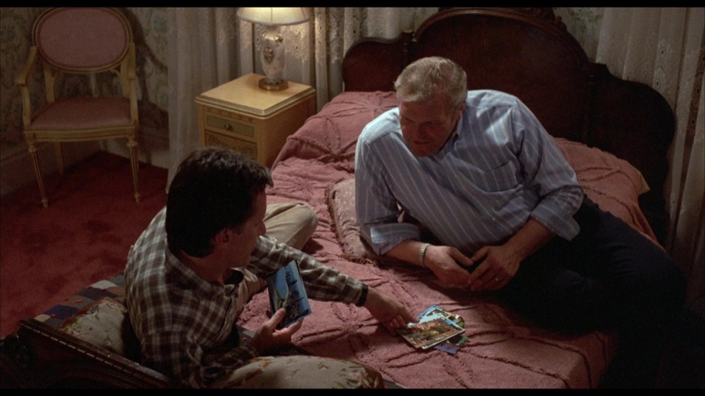Two men sitting on a bed, looking at photographs, with a lamp and a chair in the background.