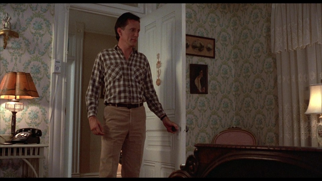 A man wearing a plaid shirt and beige pants stands at a doorway, holding the doorknob. The room features patterned wallpaper, a vintage lamp, a phone, and framed artwork.