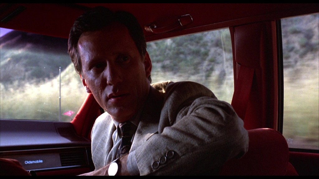 A man in a light-colored suit sits in a red car, looking intently out the window, with a serious expression on his face.