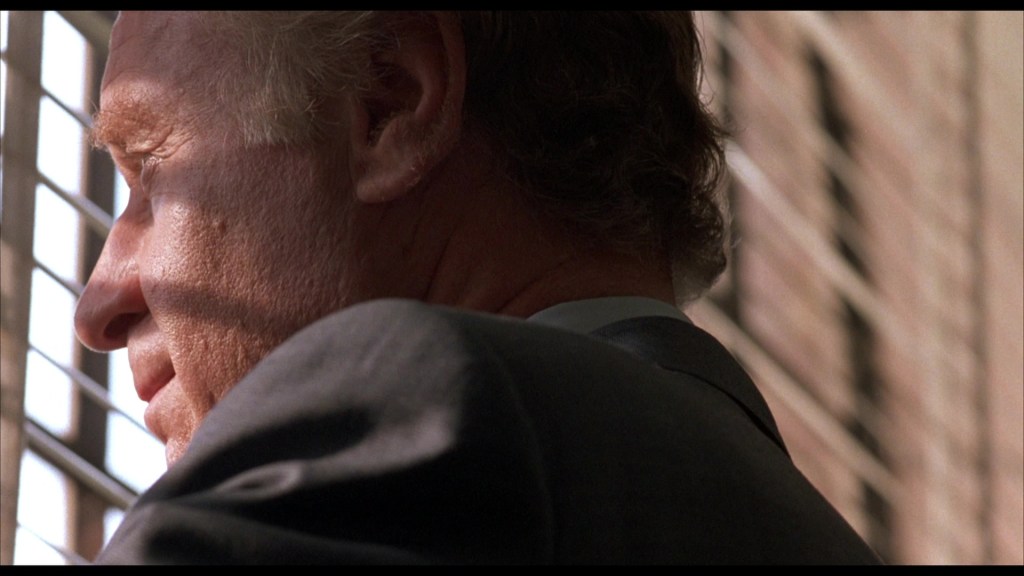 Close-up of a man in a suit looking out through window blinds, with shadows cast on his face.