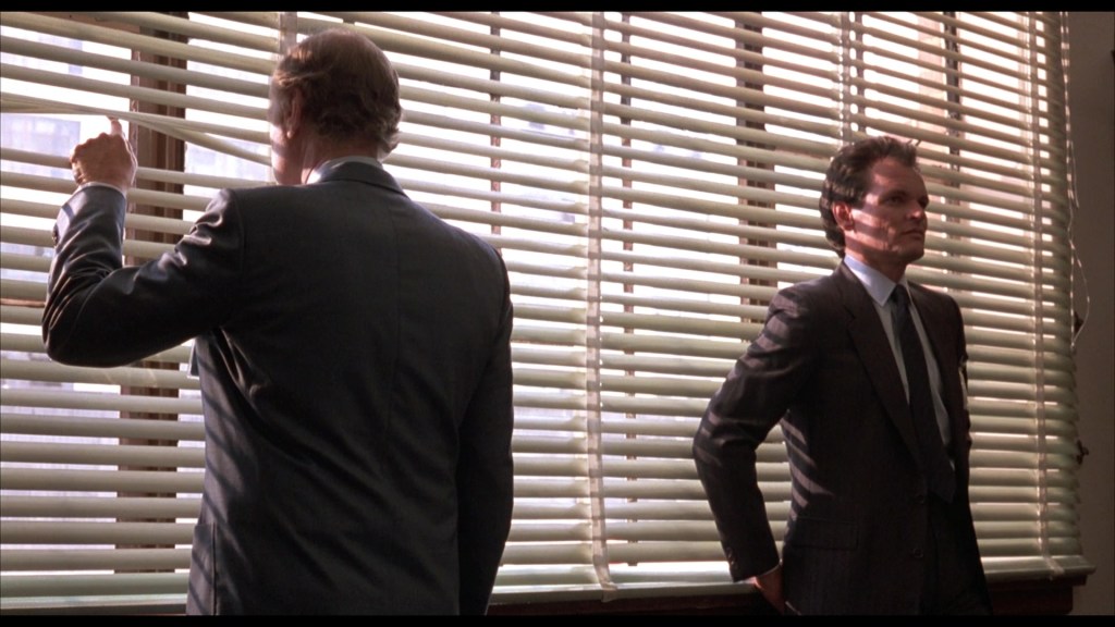 Two men in suits stand in an office with vertical blinds. One man is pulling the blinds apart to look outside, while the other stands with arms crossed, watching.