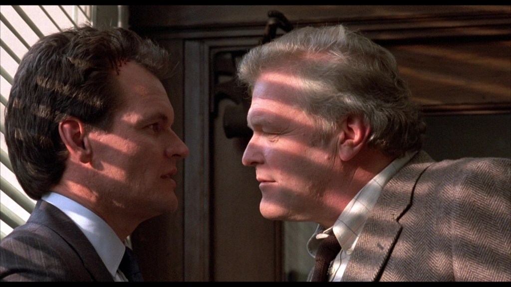 Two men facing each other closely, with intense expressions. Light and shadow create lines across their faces.