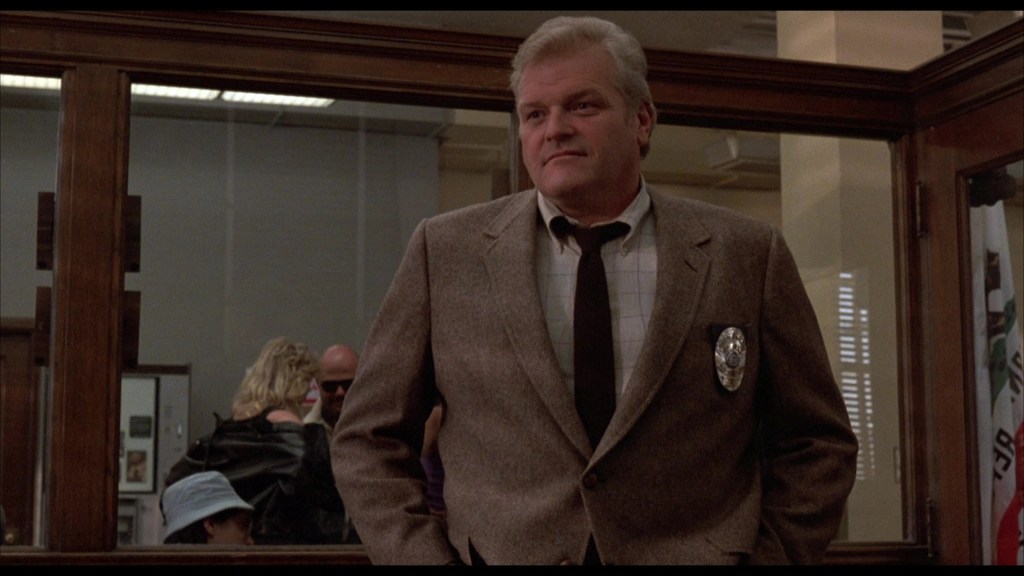 A man in a brown blazer and tie stands inside a police station, with a badge visible on his chest. He appears to be engaged in conversation while looking at the camera.