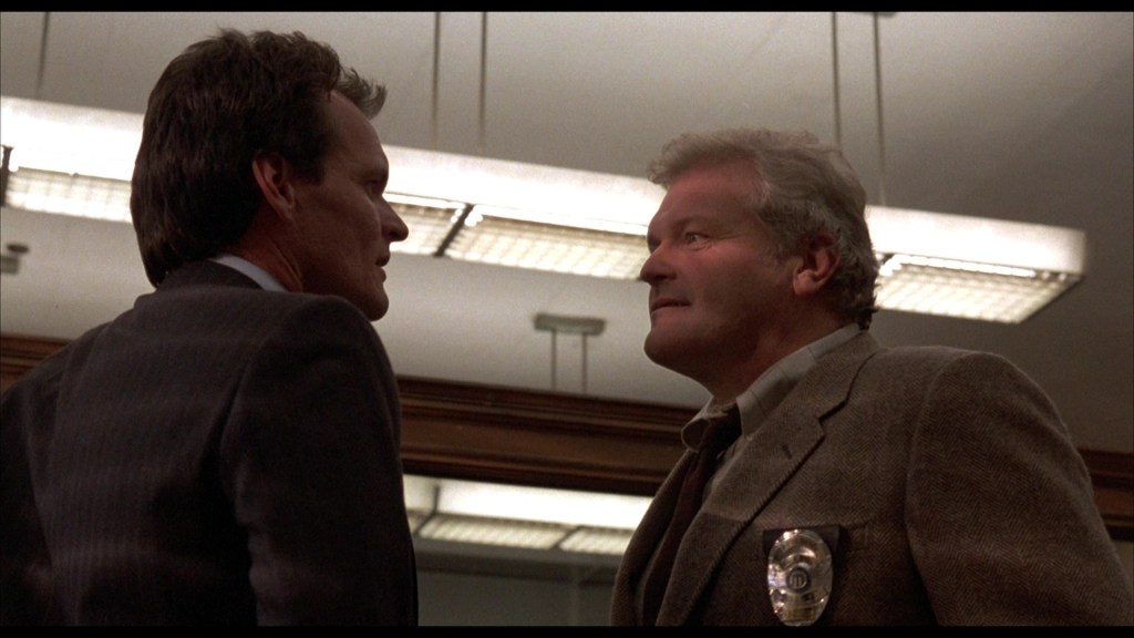 A tense confrontation between two men in an office setting, one in a suit and the other in a tweed jacket, with a police badge visible on the latter.