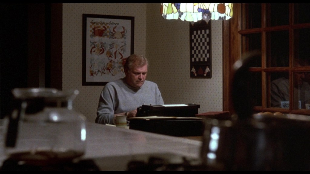 A man sits at a table typing on a typewriter in a dimly lit room, illuminated by a stained glass lamp. In the background, there are framed artworks and a chessboard on the wall.