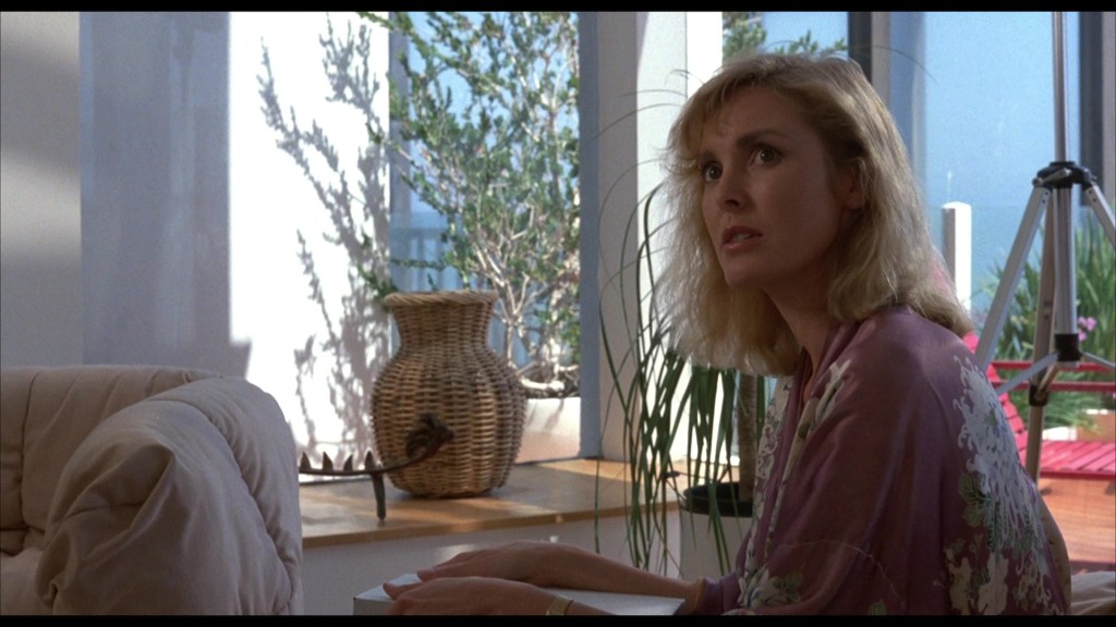 A woman with blonde hair sits in a light-filled room, looking intently with an expression of concern. She is wearing a purple robe, surrounded by indoor plants and a woven basket.