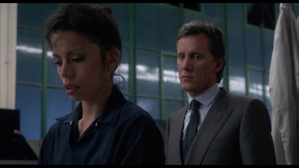 A woman in a blue shirt is looking down, focused on her work, while a man in a gray suit stands behind her, observing with a serious expression in a well-lit industrial setting.