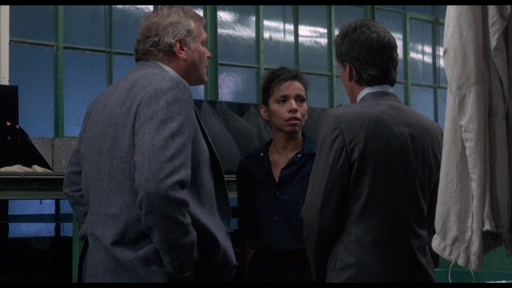 Three people engaged in conversation in a dimly lit room with green windows, two men in suits and one woman in a dark shirt.