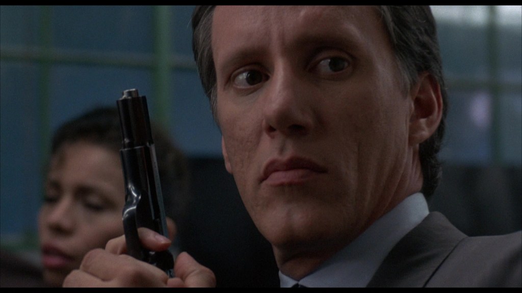A man in a suit holding a handgun, looking tense and focused, with a woman partially visible in the background.