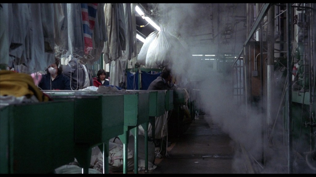Inside a laundry facility, workers are seen amongst hanging garments. Steam fills the air, creating a misty atmosphere. The setting features green tables and equipment along the sides.